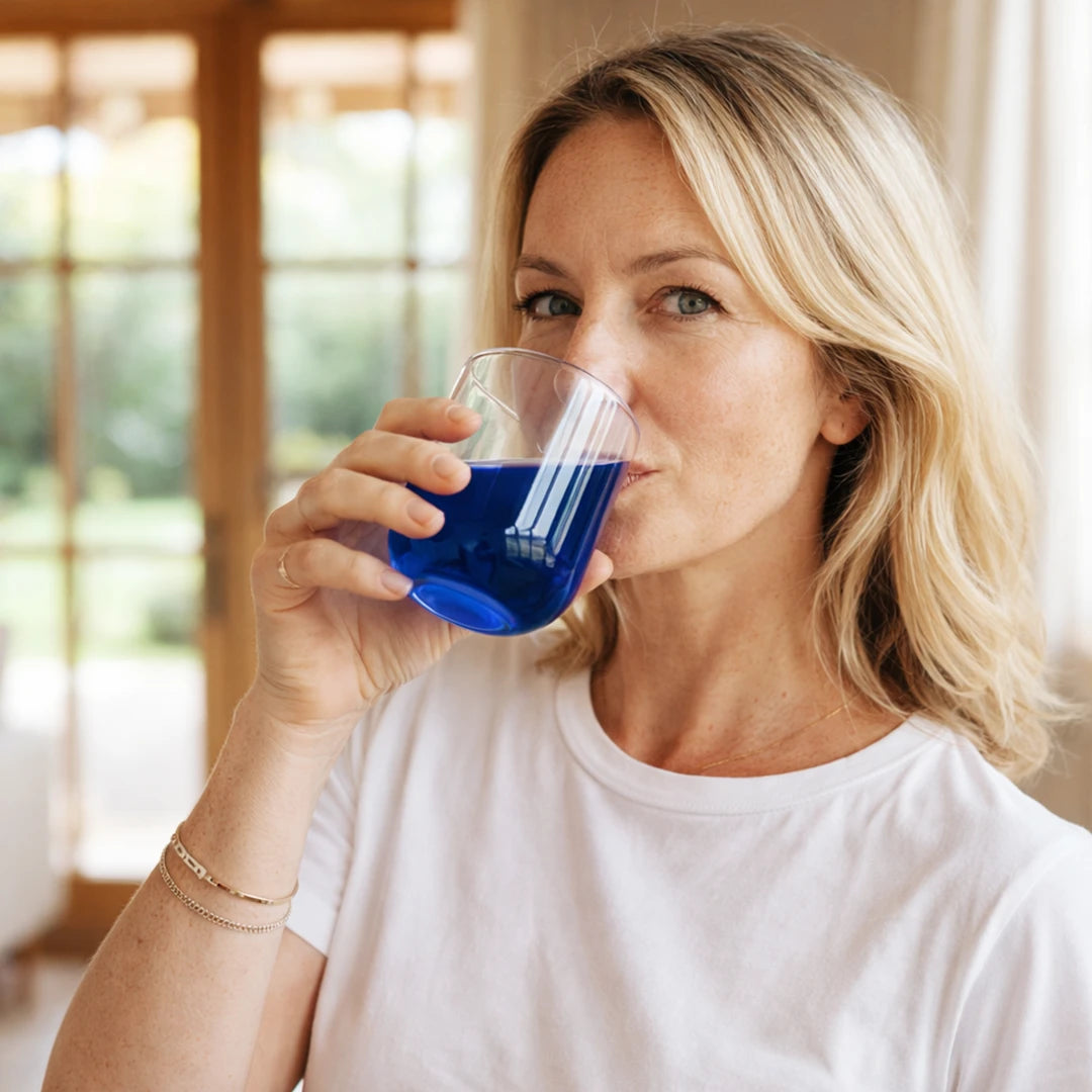 Femme d’environ 45 ans buvant une boisson bleue à base de phycocyanine dans un verre, dans un intérieur lumineux avec grandes fenêtres et ambiance lifestyle bien-être.